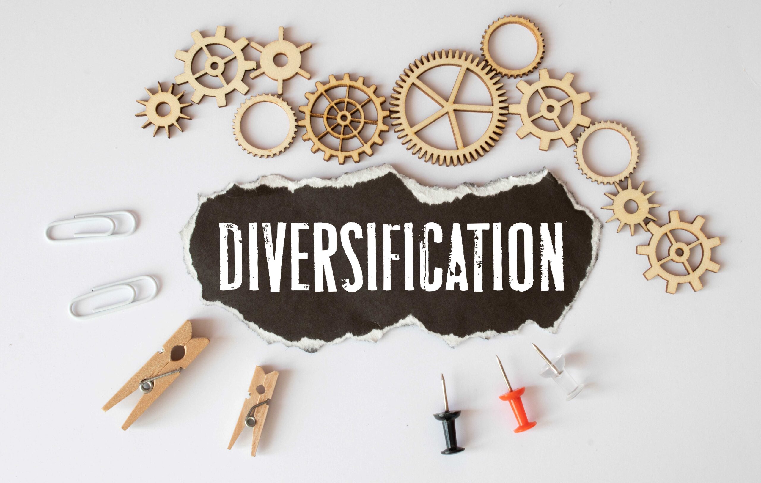 Graphic of a ripped piece of paper with the word 'Diversification' showing through alongside wooden cogs, pins, and pegs - portfolio diversification